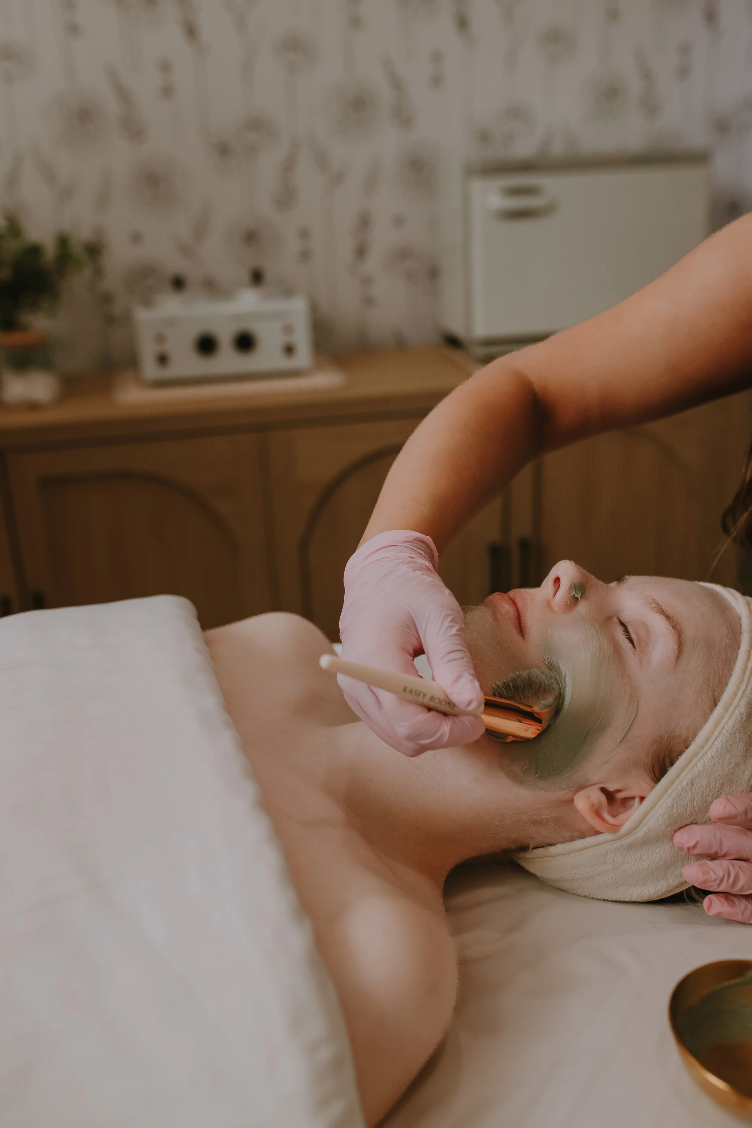 A person lying on a bed getting a facial treatment with a green facial mask, while someone else applies a mask with a brush, in a cozy room with vintage wallpaper and a cabinet in the background.