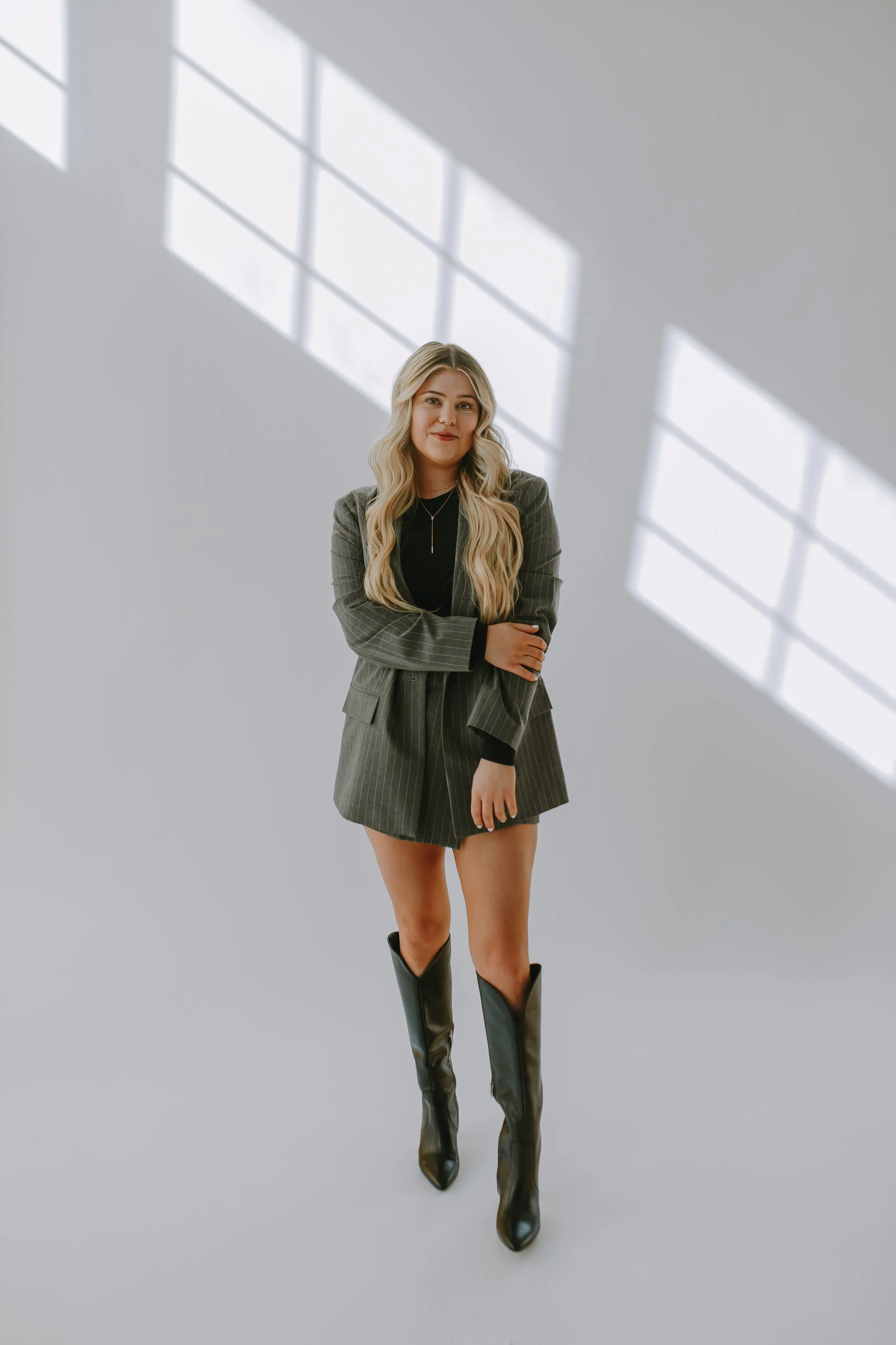 A young woman with long blonde hair standing in a minimalistic room with large windows and natural light. She is wearing a dark top with a plaid blazer, matching plaid shorts, and black knee-high boots.