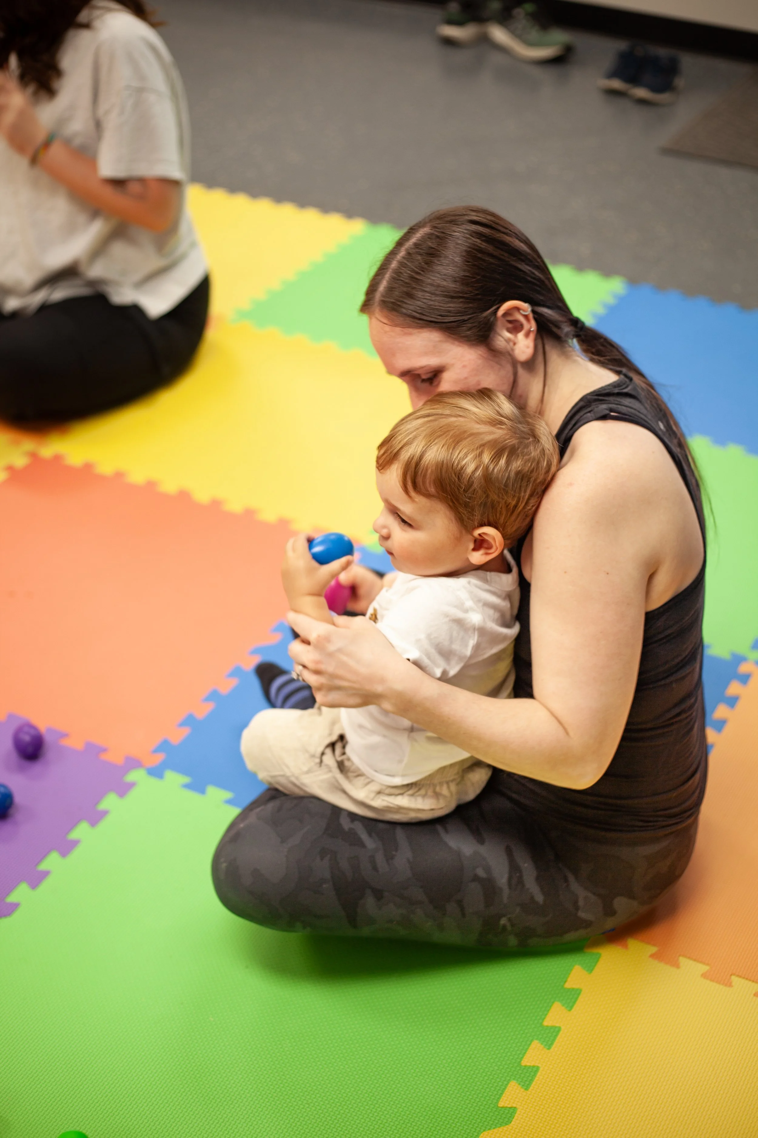 A woman and young child sitting on colorful foam mat in a play area, the woman holding the child and both engaging in play with small, colorful egg shakers.