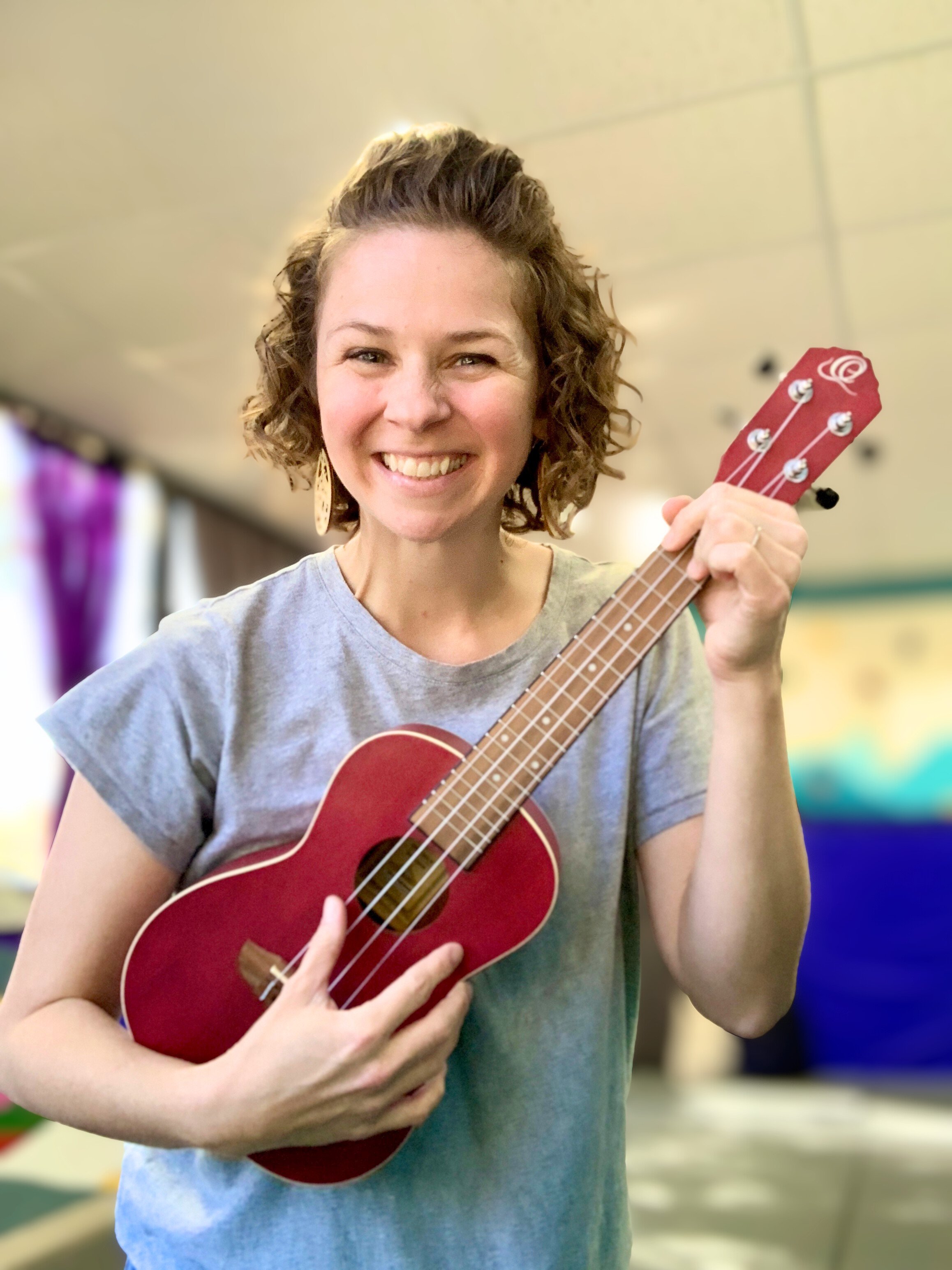 A woman with curly hair smiling and holding a small red ukulele.