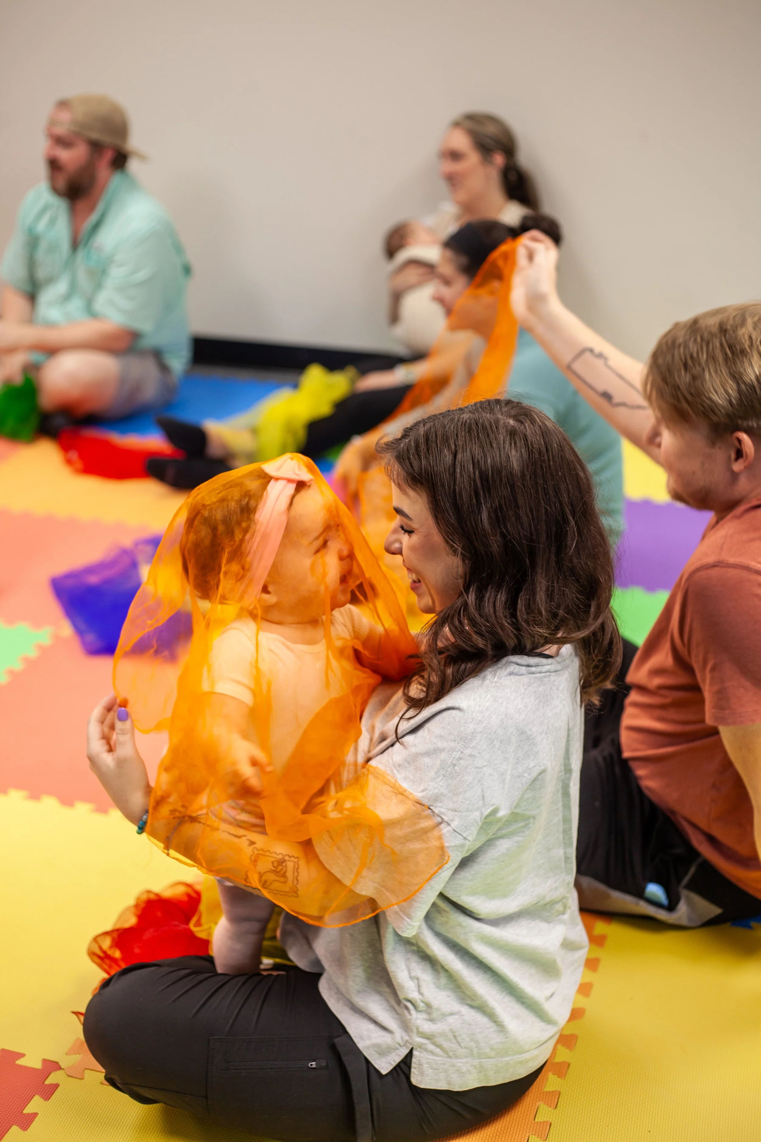 A group of people sit on the colorful foam mats on the floor, with some engaging in a sensory activity involving orange silk scarves, in a child-friendly room.
