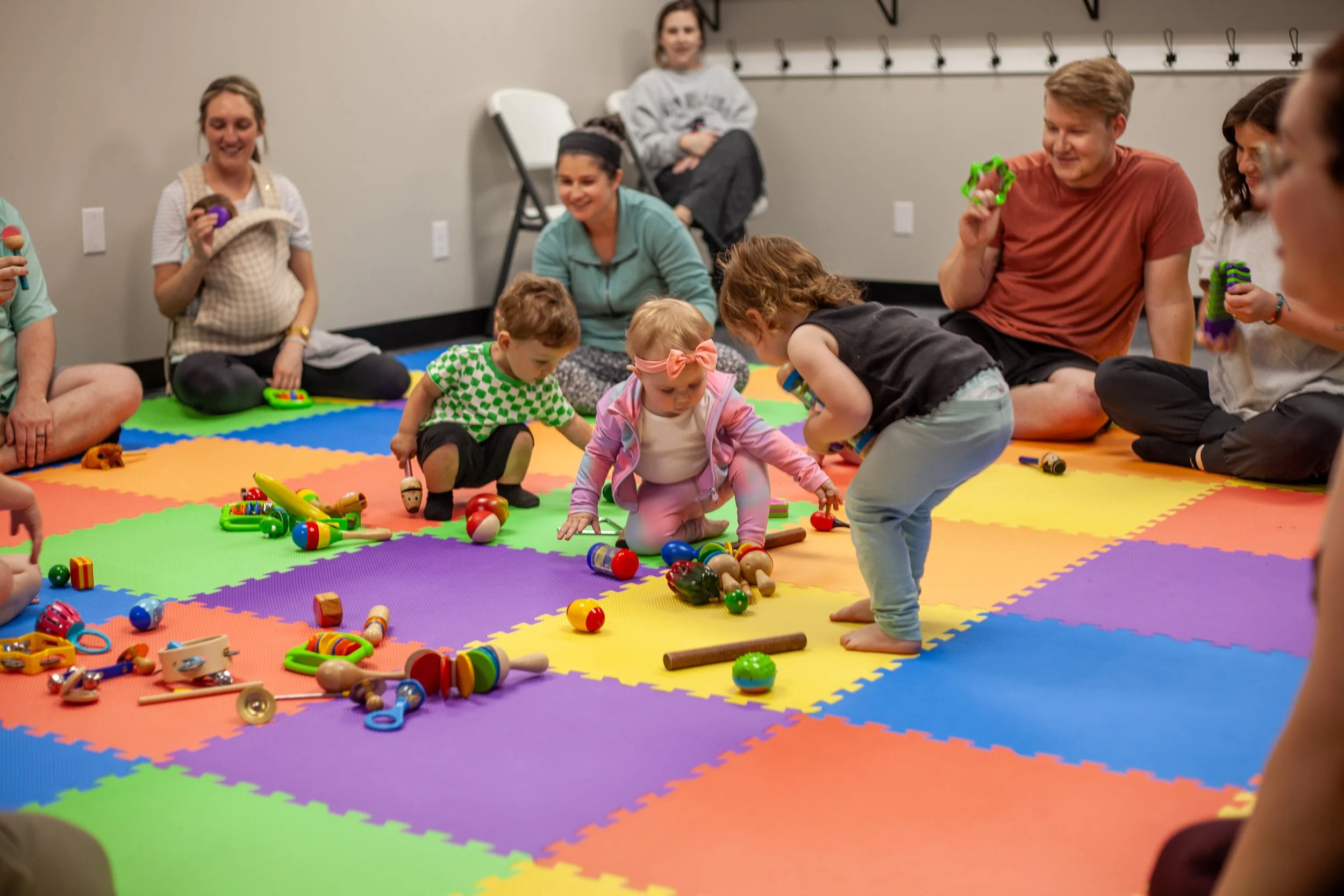Children and adults sitting on colorful foam mats playing with toy maracas, shakers, and other instruments in a room with beige walls and hooks on the wall.