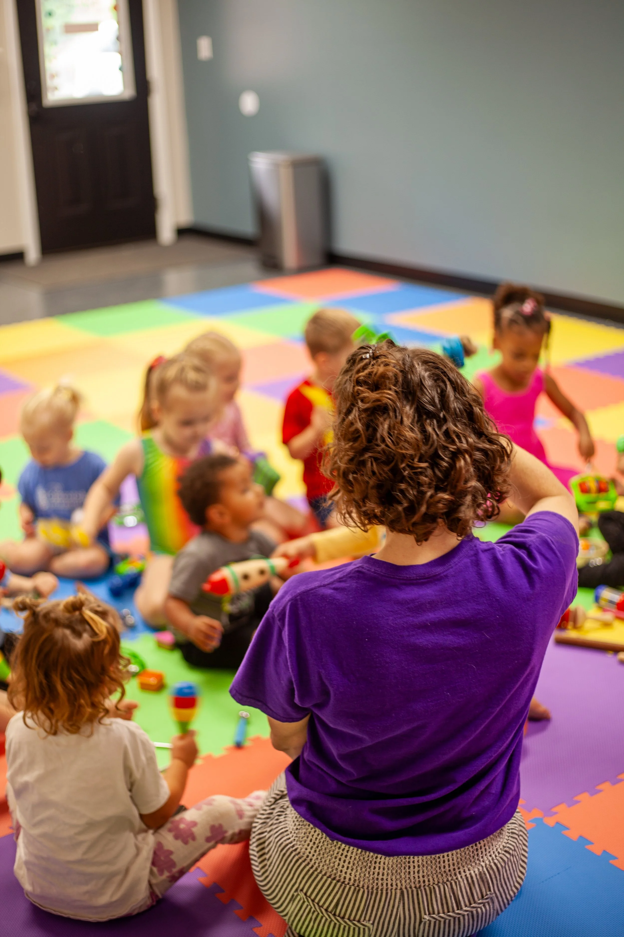 A group of young children and an adult sitting on multicolored foam mats in a room with a blue wall and a black door, engaging in a play activity with instruments.