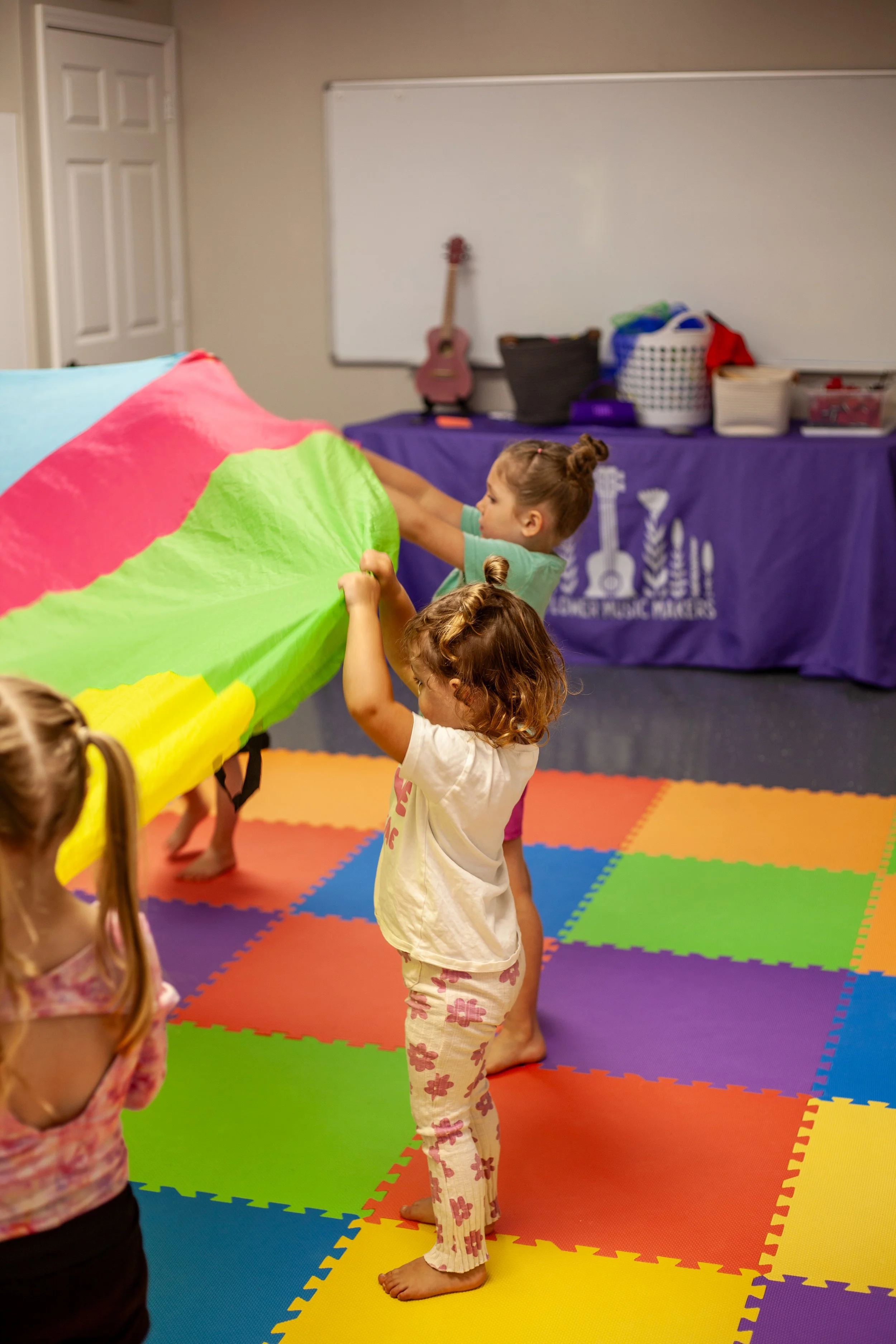 Young children in activewear playing with a colorful parachute in a room with a rainbow-colored foam mat, in front of a purple table with various baskets and a guitar leaning against a whiteboard.