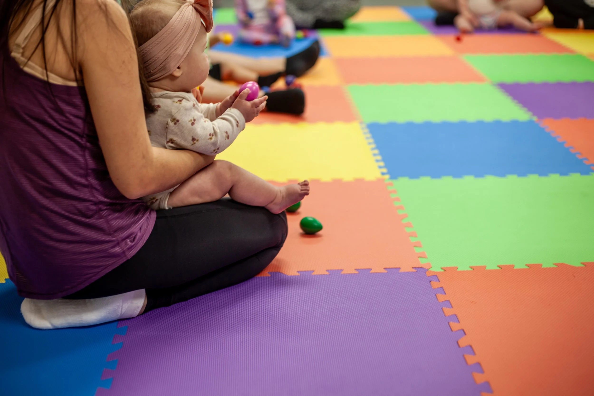 A young child sitting cross-legged on a colorful foam play mat, holding a pink plastic shaker egg, with an adult sitting behind her on the mat, and other children in the background. The scene appears to be a children's activity or play area.