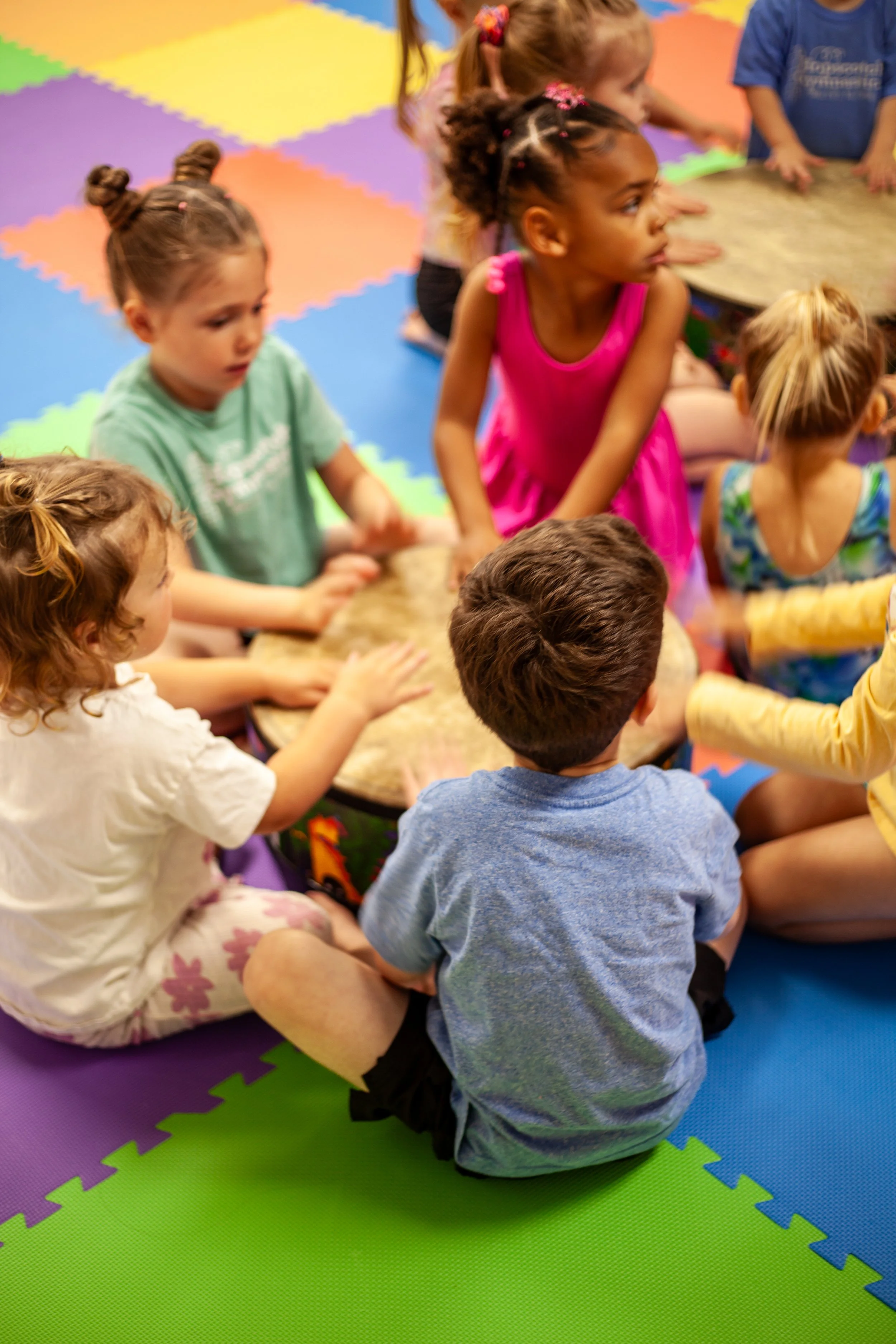 Children seated on colorful interlocking foam mats, gathered around a drum, engaging in a music activity.