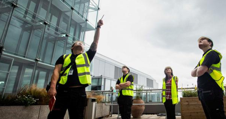 Four construction workers in high-visibility vests standing outside a modern glass building, with one pointing upwards.