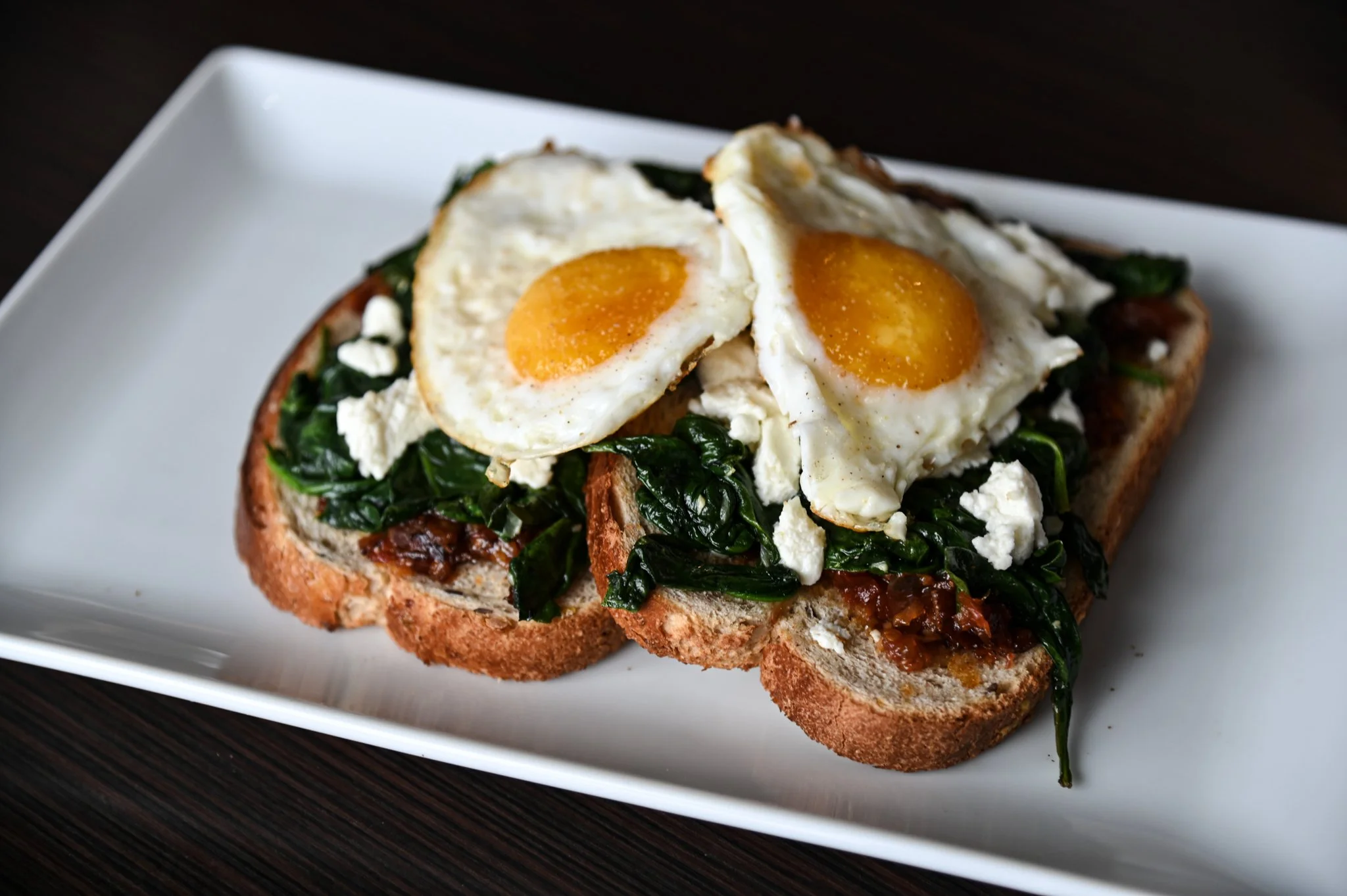 Open-faced toast with sautéed spinach, over-easy eggs, and melted cheese on a white plate.