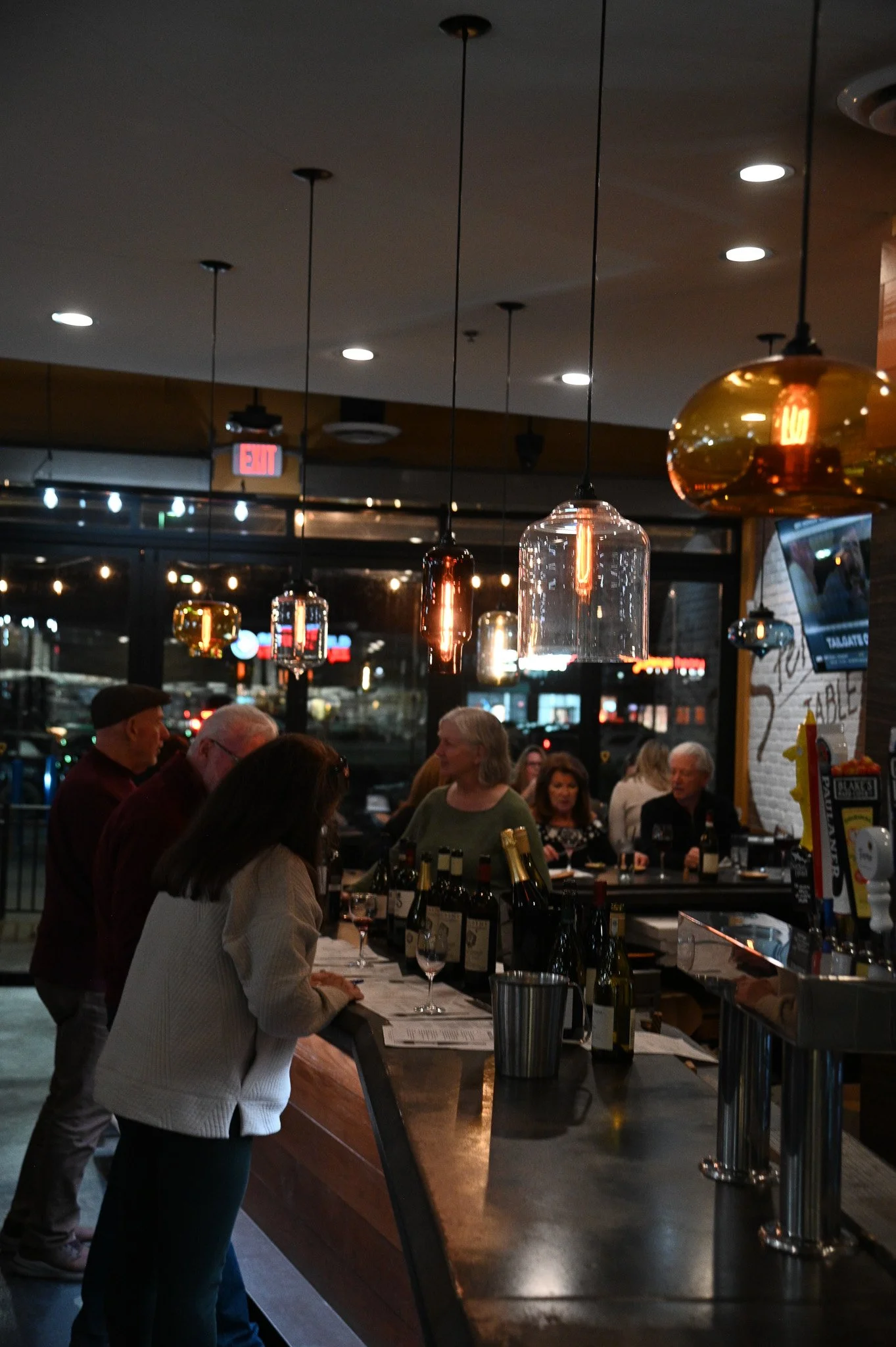 People ordering and serving drinks at a bar inside a restaurant at night, with hanging pendant lights and a crowded background.