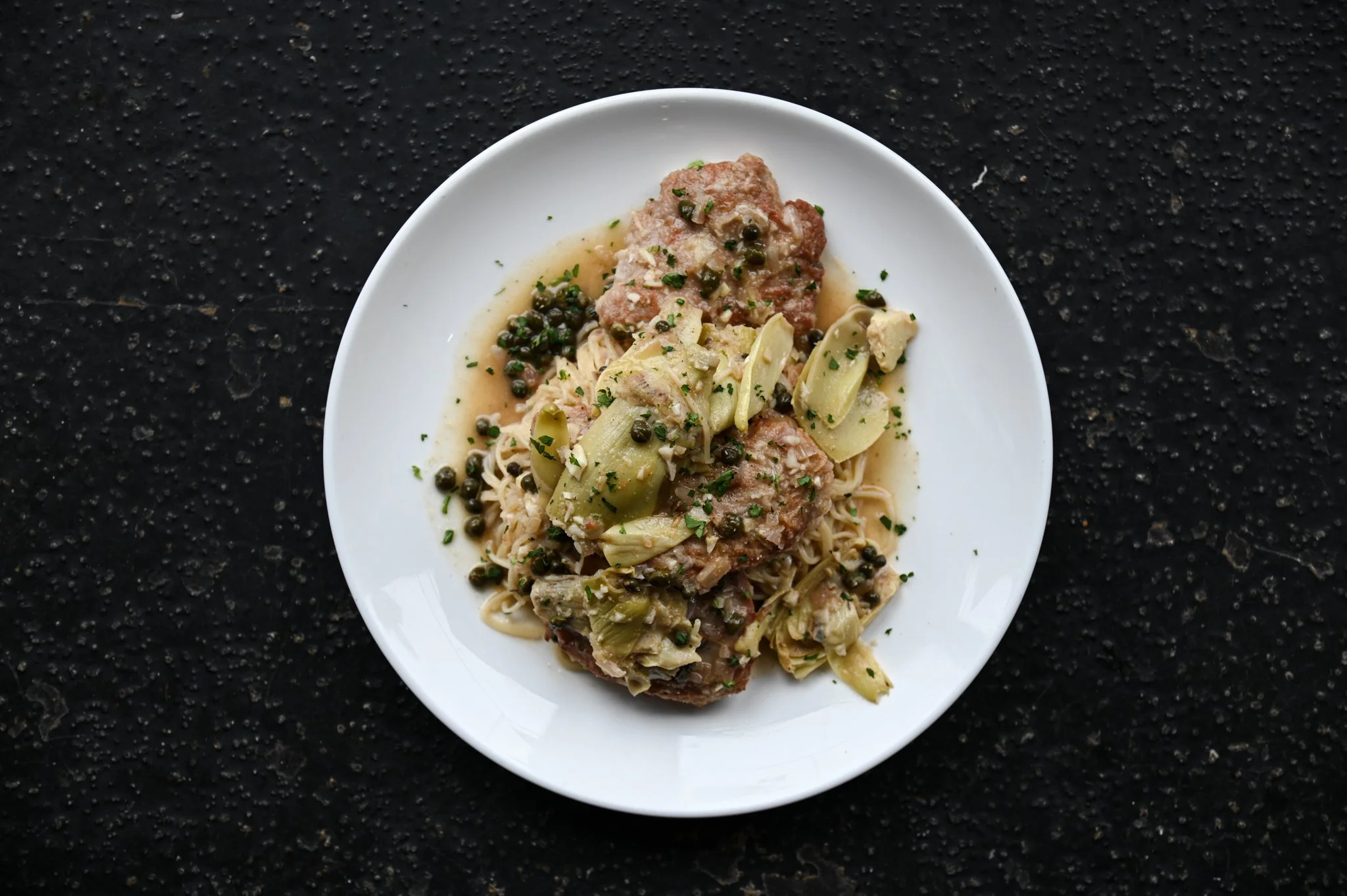 A white plate of pasta with chicken, green olives, and herbs on a black textured surface.