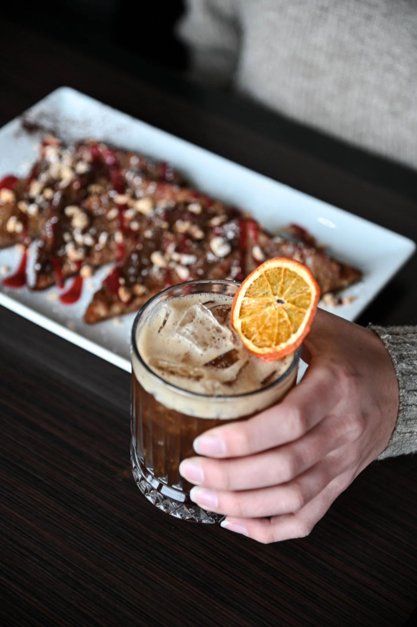 A hand holding a glass of iced beverage with a slice of dried orange on top. In the background, there is a white rectangular plate with a dessert topped with strawberries, nuts, and syrup.