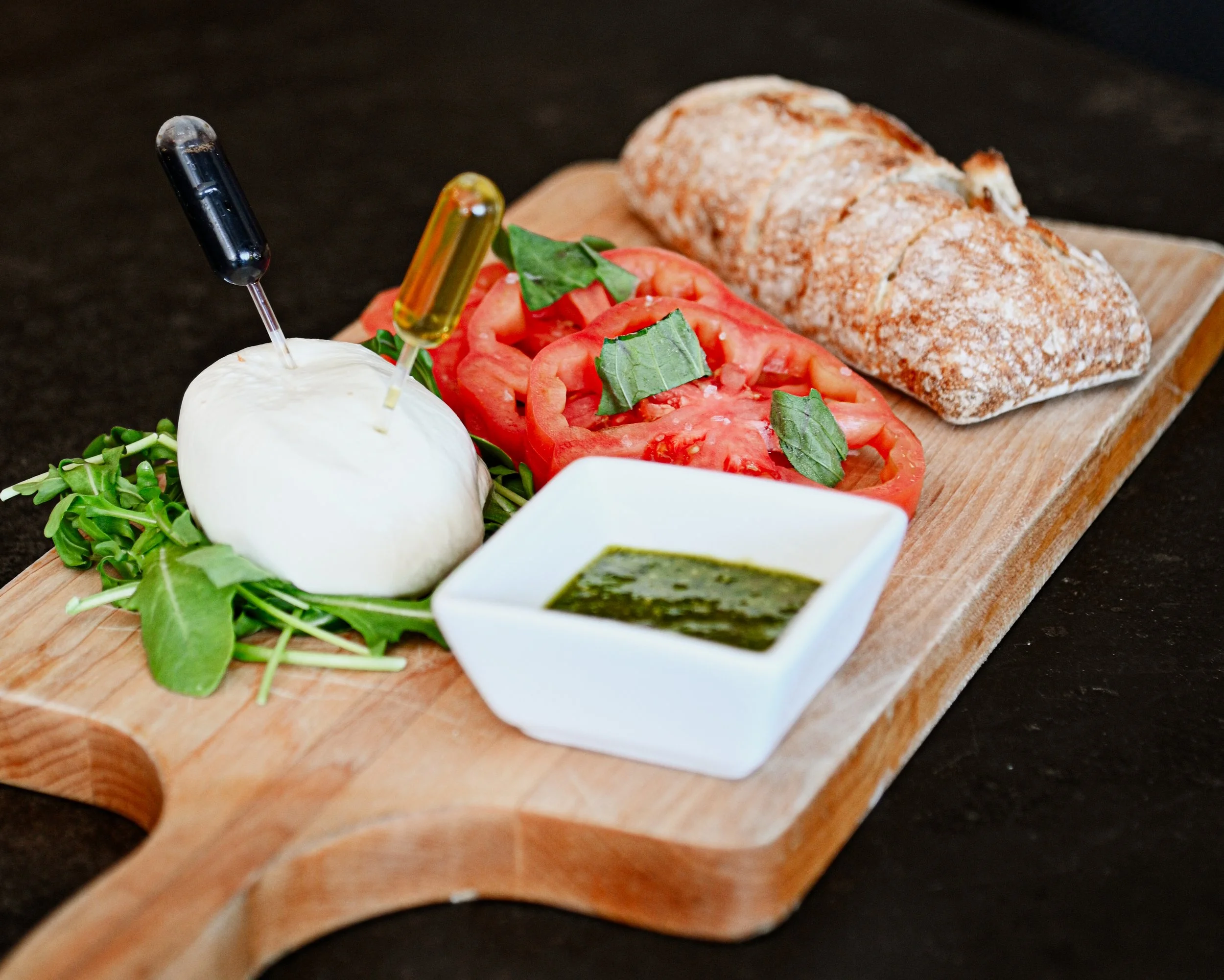 Wooden serving board with sliced tomatoes, fresh basil, two rustic bread rolls, a ball of burrata cheese, a small dish of green pesto, and two oil capsules, on a dark surface.