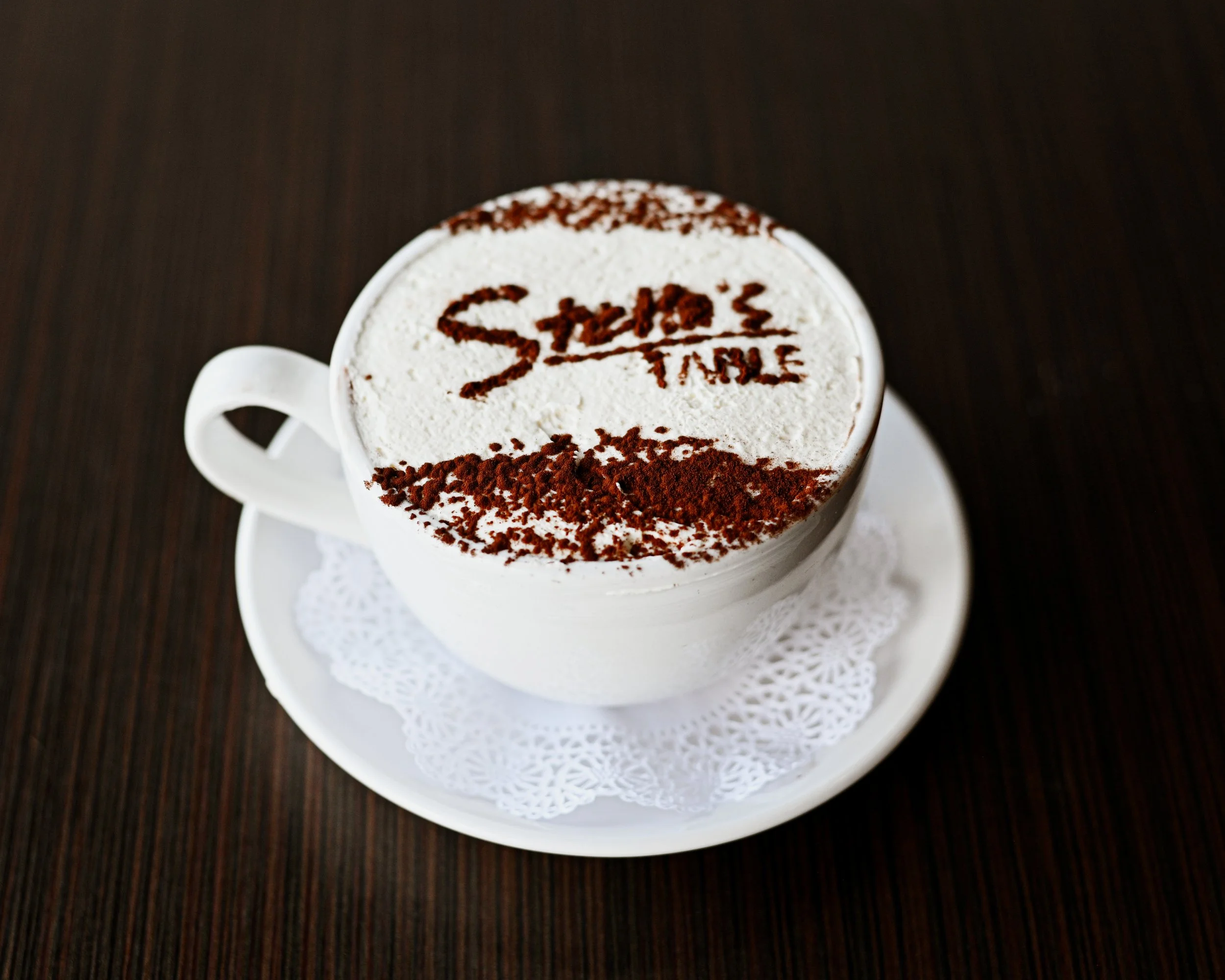 A white coffee cup filled with a foamy beverage, topped with whipped cream, cocoa powder, and the words 'Stay Time' written in cocoa powder on top, placed on a white saucer with a paper doily, on a dark wooden surface.