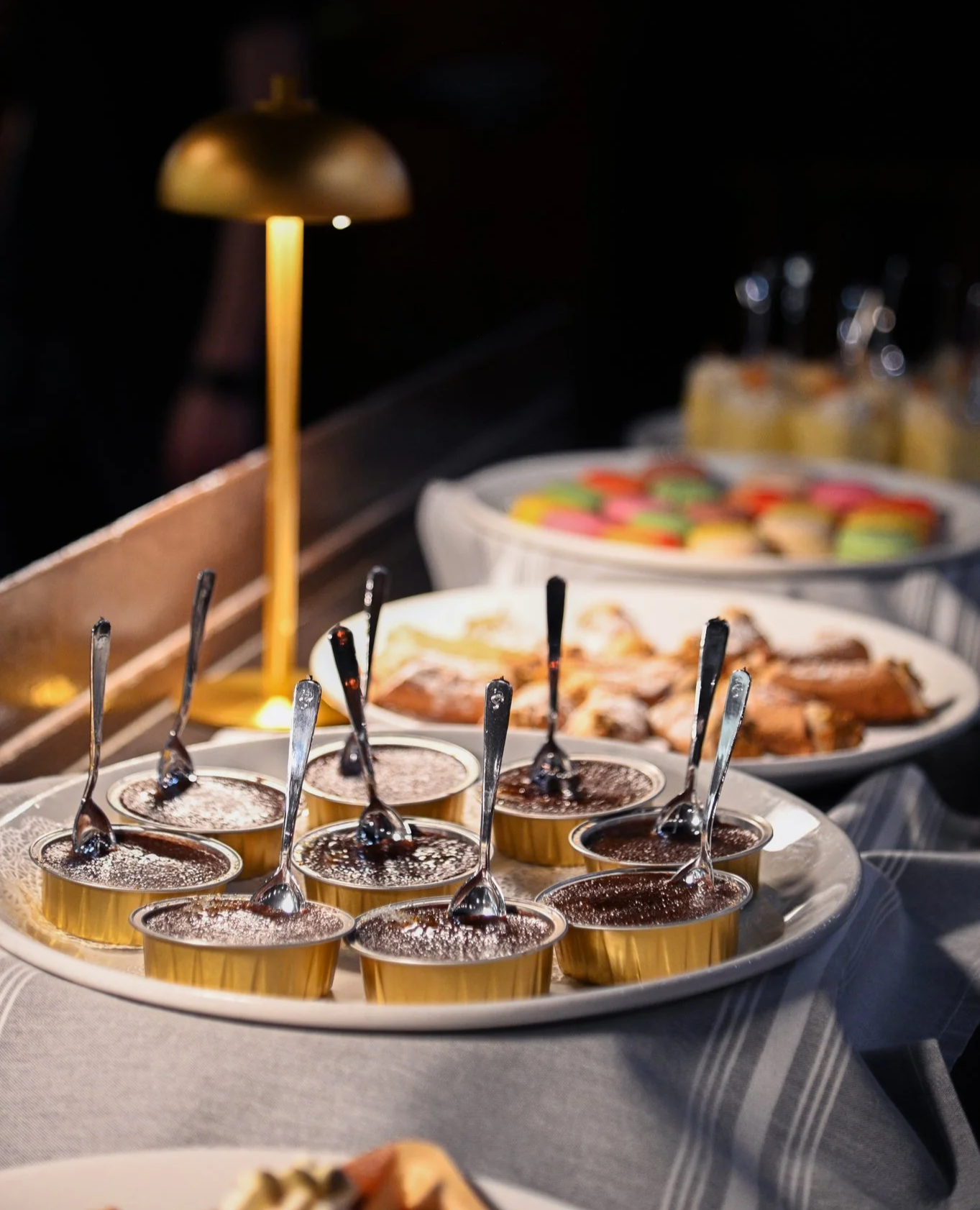 Dessert table featuring small chocolate puddings in gold cups with spoons, a platter of decorated cookies, and a plate of sliced meat or fish. A lamp provides soft lighting in the background.