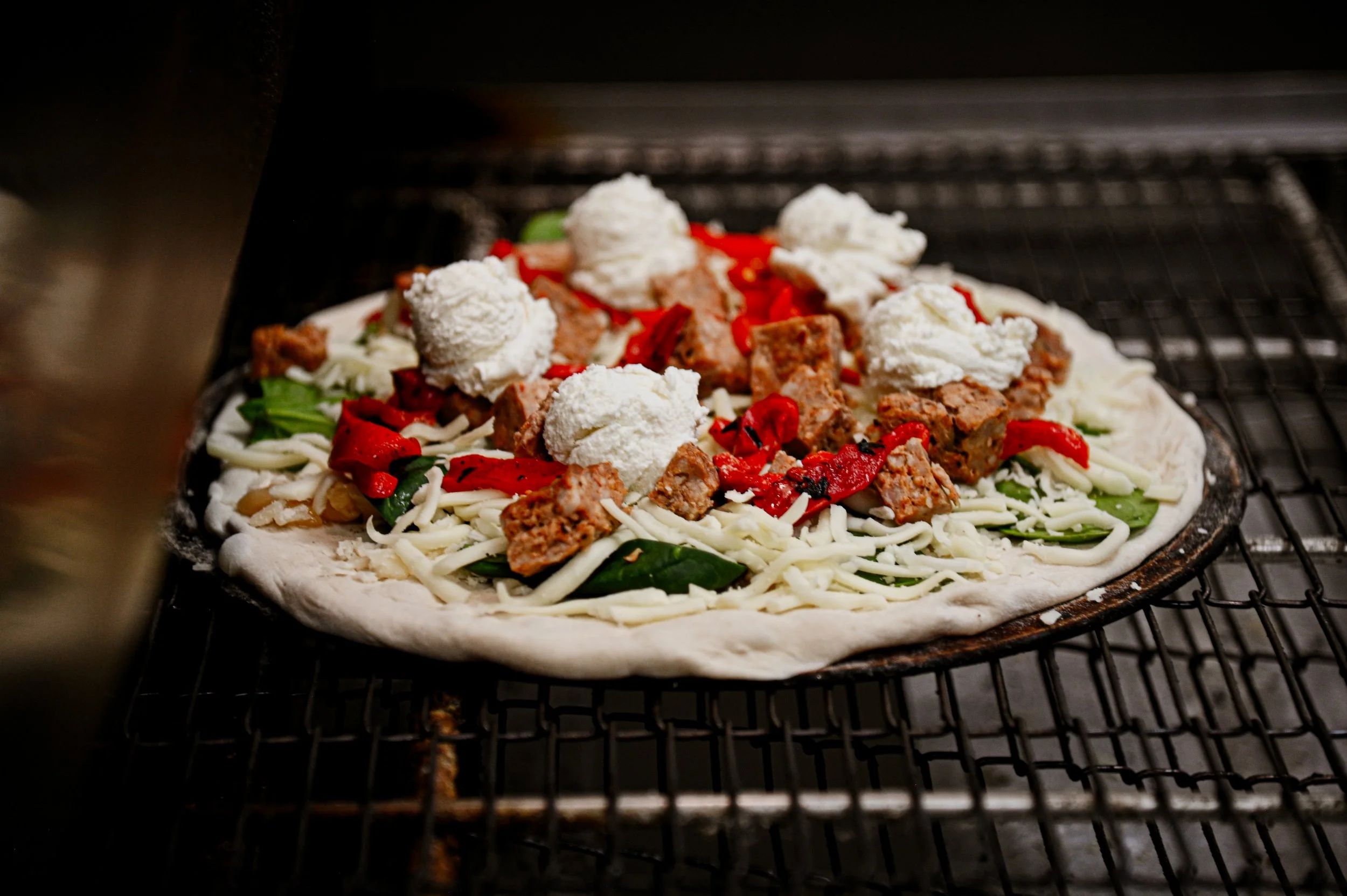 A pizza with cheese, spinach, red peppers, meat, and dollops of ricotta cheese is baking in an oven on a wire rack.