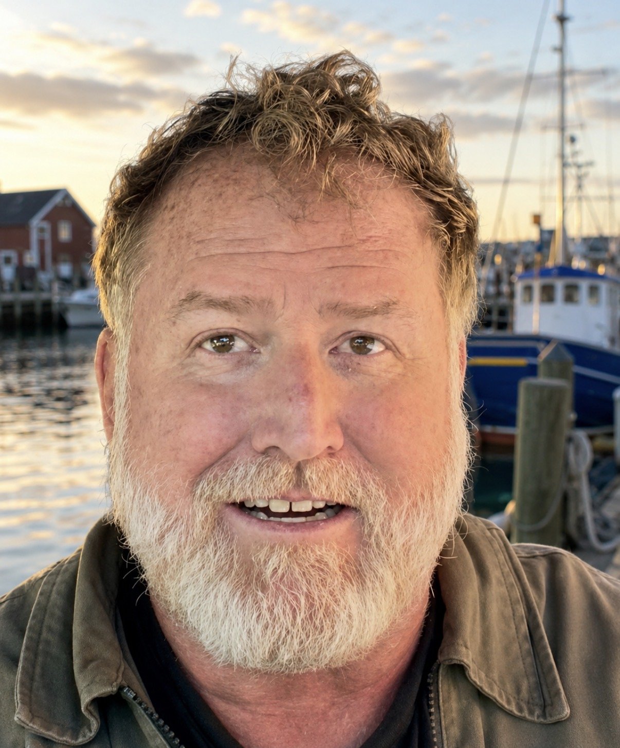 A middle-aged man with curly hair and a beard smiling outdoors near a marina with boats, during sunset.