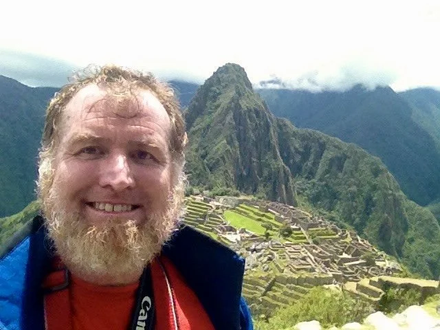 Smiling man with beard in front of Machu Picchu and Huayna Picchu mountain in Peru.