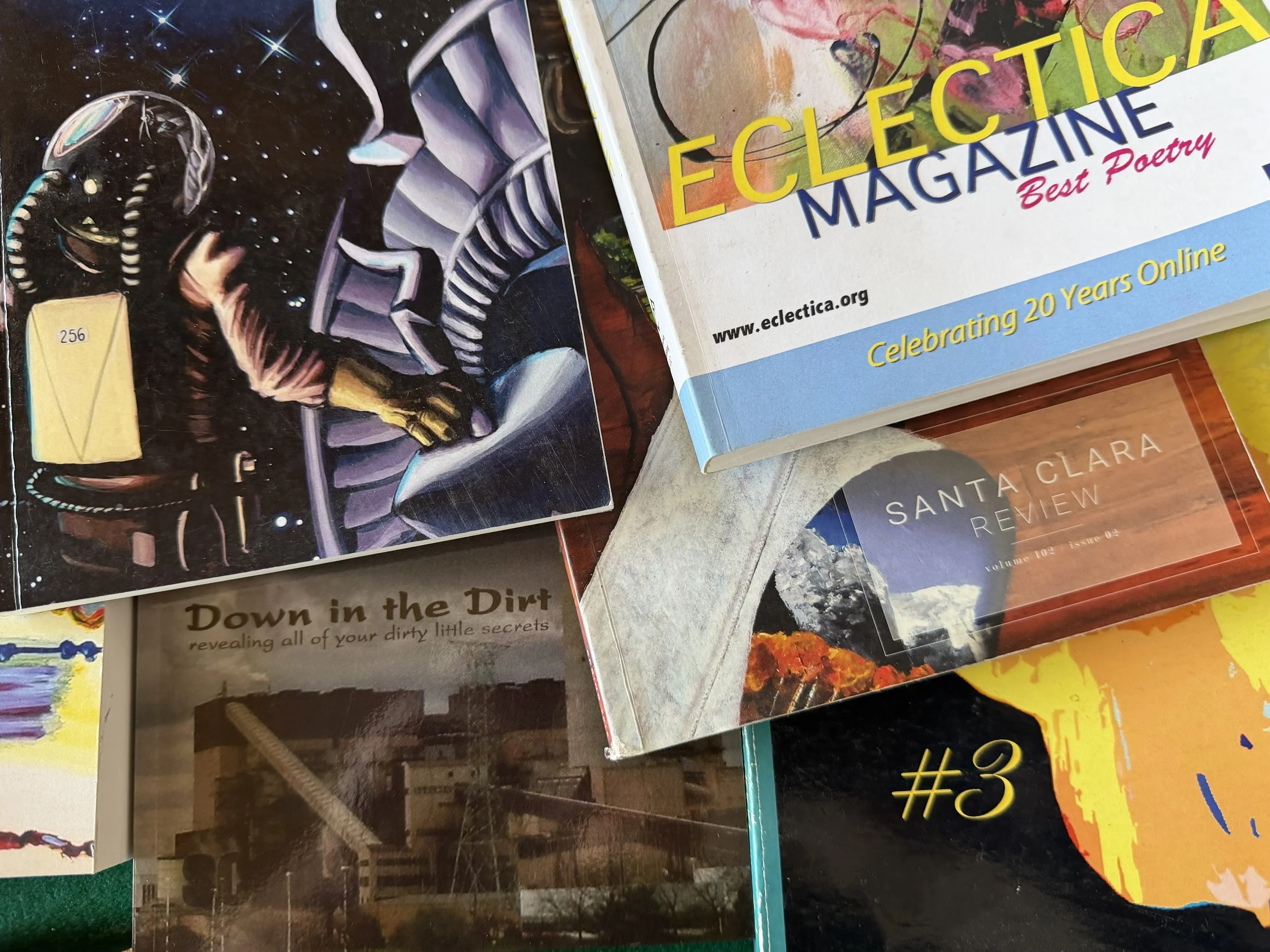 Several magazines and booklets stacked together on a surface, including a magazine titled "ELECTIC MAGAZINE" with a colorful cover and the phrase "Celebrating 20 Years Online." Other visible titles include "Down in the Dirt" and "Santa Clara Review."