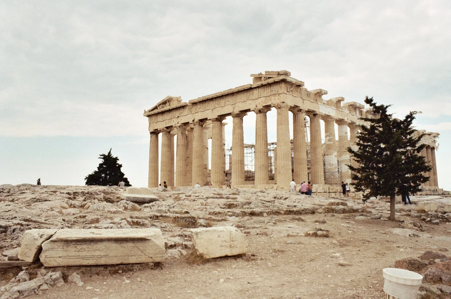 Ancient Greek Parthenon temple on the Acropolis with several visitors and sparse trees in a rocky landscape.