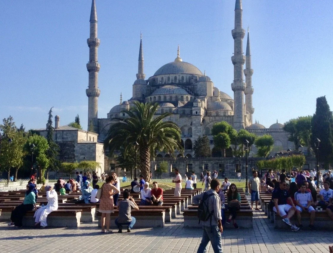 People sitting and walking in a park with a large historic mosque, Sultan Ahmed Mosque (Blue Mosque), in the background with multiple domes and minarets under a clear blue sky.