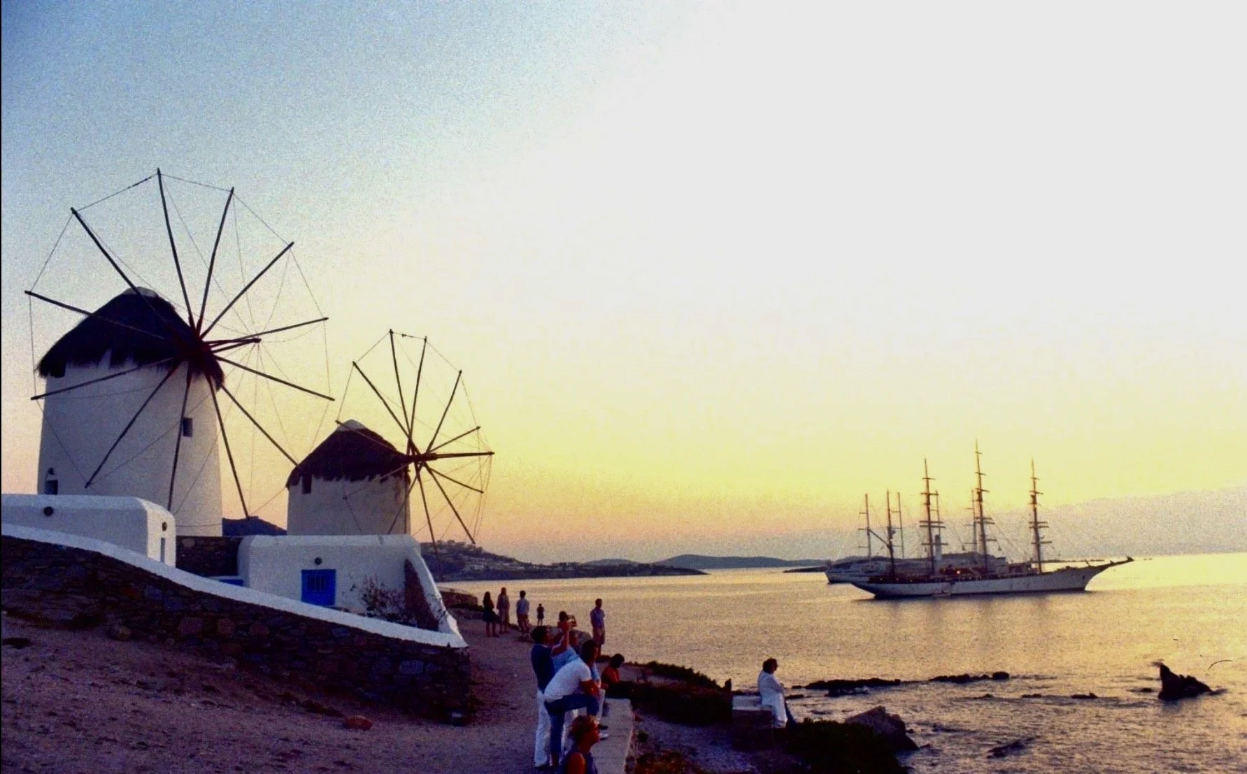 Windmills with sails, a sailing ship on water, people walking along the shoreline during sunset.