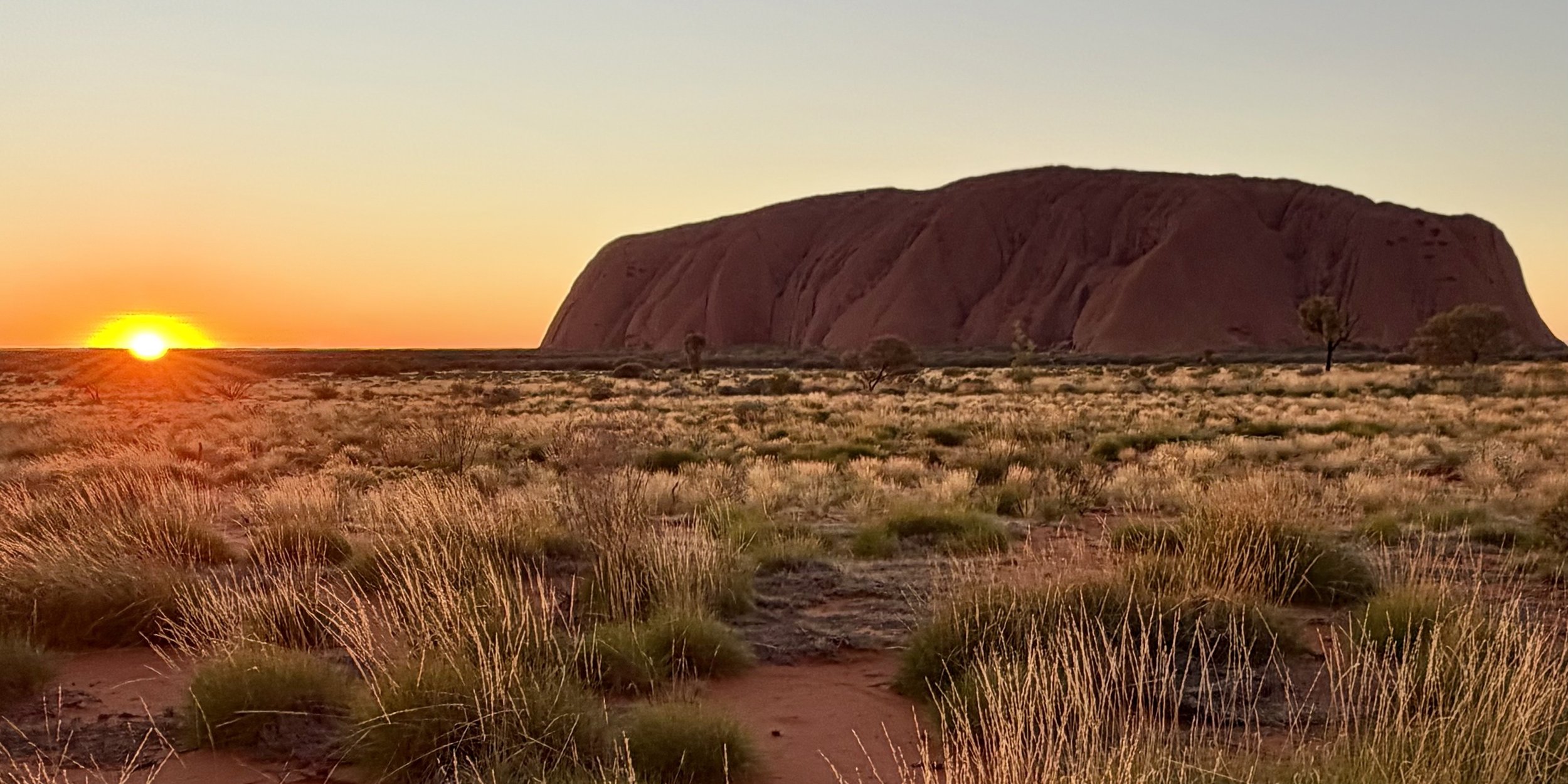 Sunset over a desert landscape with a large red rock formation and sparse vegetation.