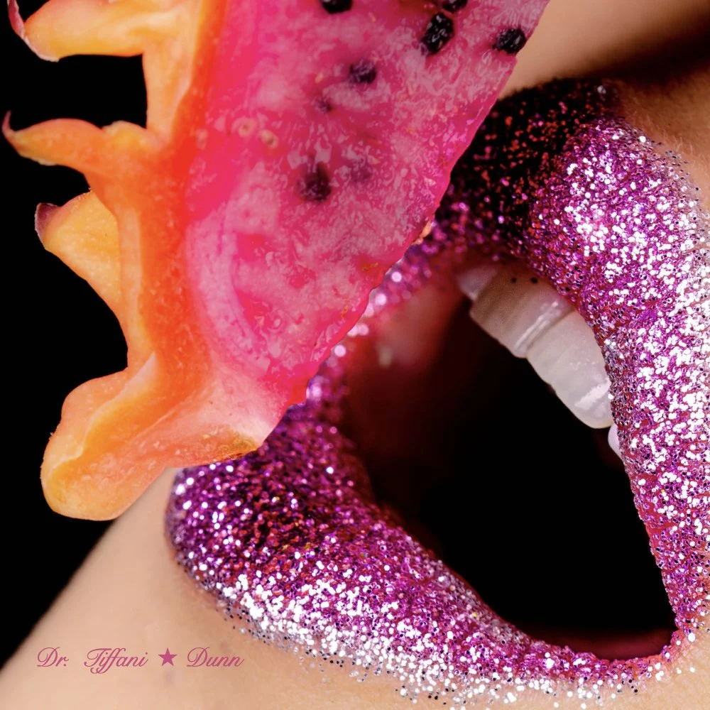 Close-up of a mouth with pink glittery lips, holding a slice of orange starfruit near the lips. The background is black.