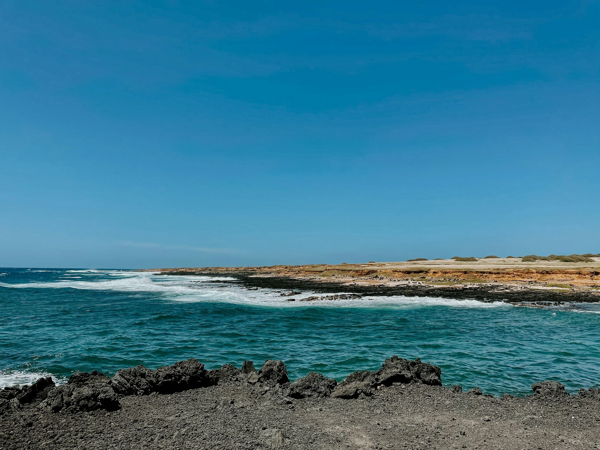 Coastal landscape with volcanic rocks in the foreground, waves crashing on the rocks, sandy shoreline, and a blue sky.