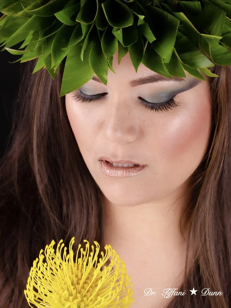 A woman with long brown hair wearing a leafy green crown, holding a yellow flower, with closed eyes and natural makeup.