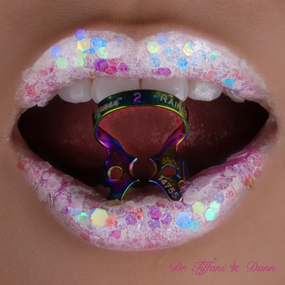 Close-up of lips with iridescent lip gloss and glitter, featuring a tongue piercing with a rainbow-colored ring. The ring displays text reading '2 RAINBOW' and has an intricate design. The bottom right corner has pink text saying 'Dr. Tiffani Dunn'.