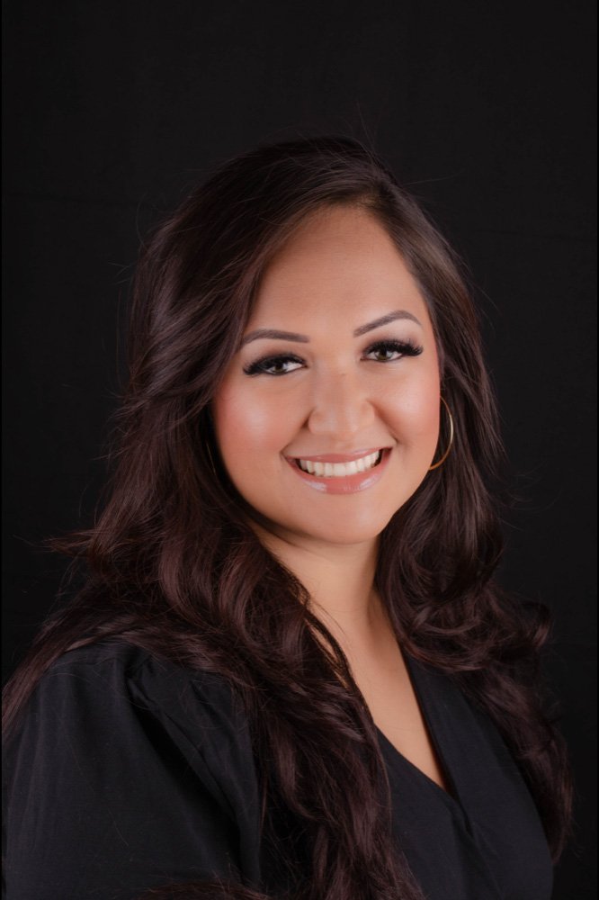 A woman with long, dark brown hair, smiling with makeup, gold hoop earrings, and a black top or dress, against a black background.