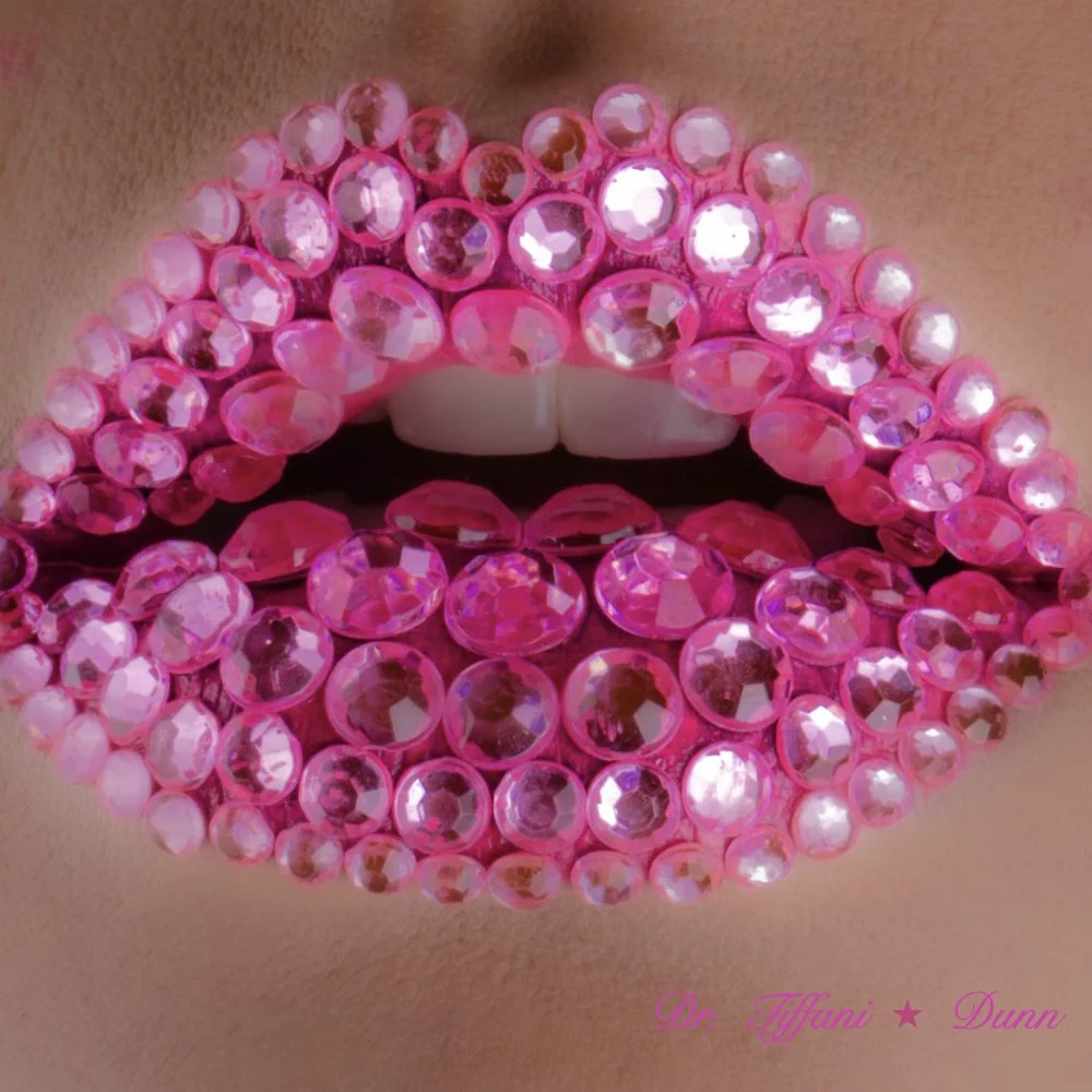Close-up of lips decorated with numerous pink rhinestones and pearls, wearing white lipstick and white teeth. Text in pink at the bottom reads 'Dr. Tiffani Duna' with a star.