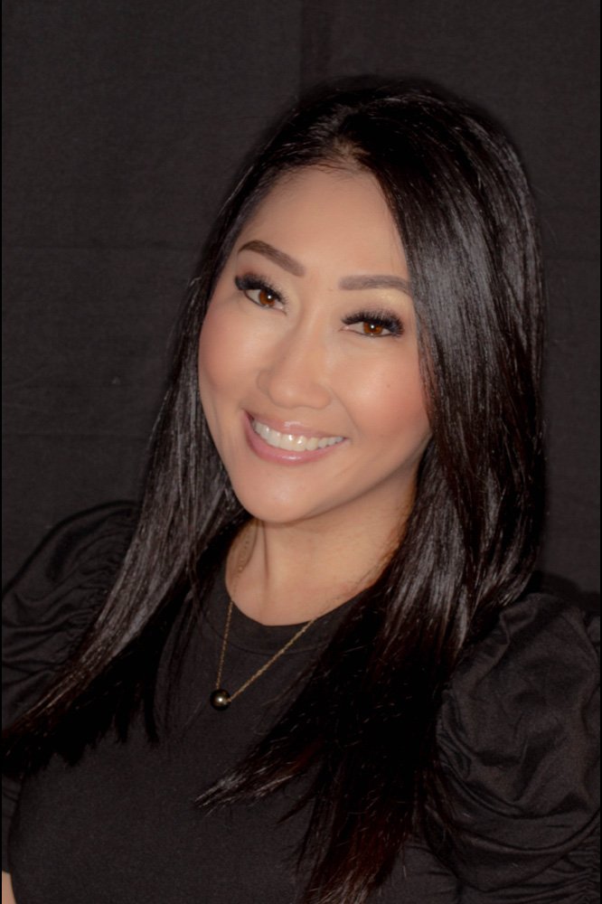 A woman with long dark hair, makeup, and a black top, smiling at the camera against a dark background.