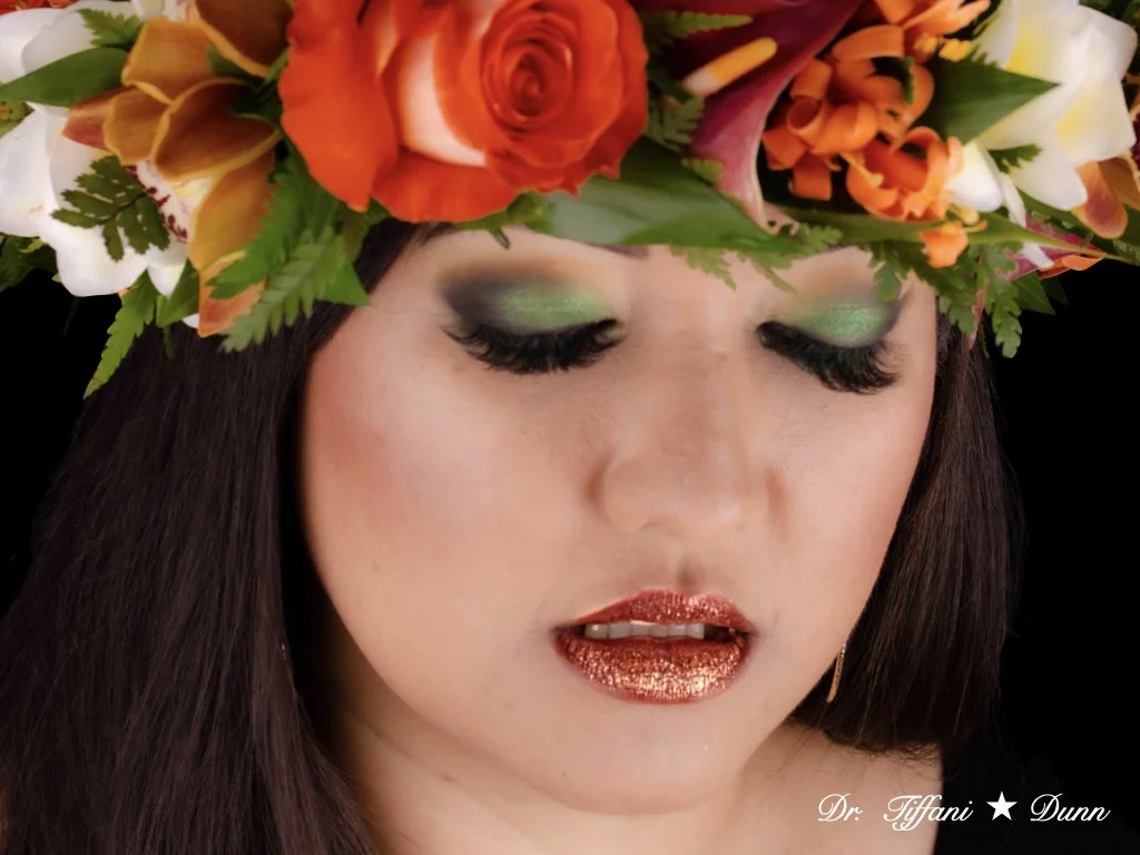 A woman with dark hair wearing a large floral crown with orange roses, yellow calla lilies, and greenery, with green eyeshadow, long black lashes, and glittery bronze lipstick.