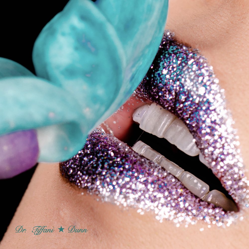 Close-up of lips with purple and silver glitter lipstick, showing white teeth, and a hand with a turquoise glove near the lips.