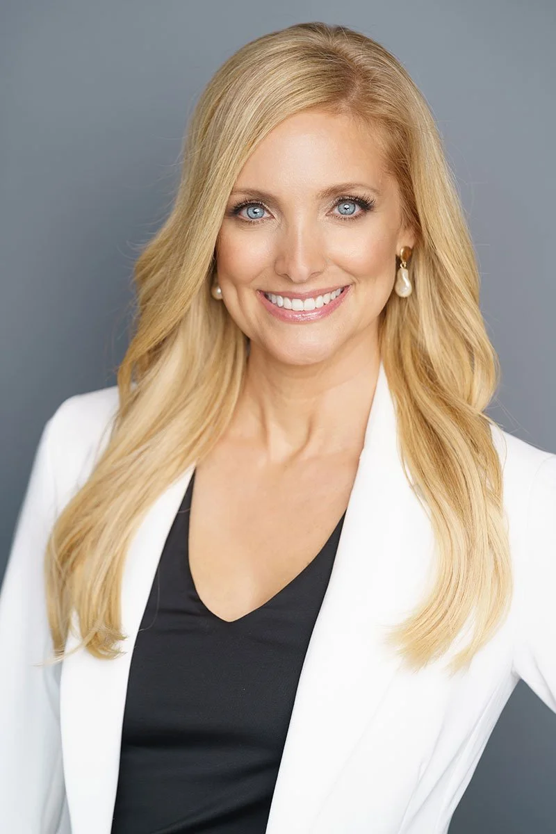 A blonde woman with blue eyes smiling, wearing a white blazer, black top, and pearl earrings, against a gray background.