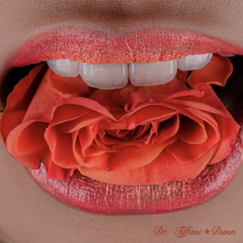 Close-up of lips painted with red lipstick, holding an orange and red rose between the lips, with the teeth showing above the rose, and the text "Dr. Tiffani★Dunn" at the bottom right corner.