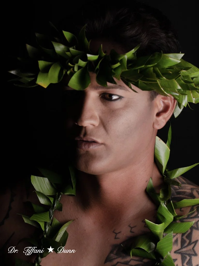 A man with dark hair and tattoos on his shoulder, wearing a leafy crown, partially covered in shadow, against a black background.