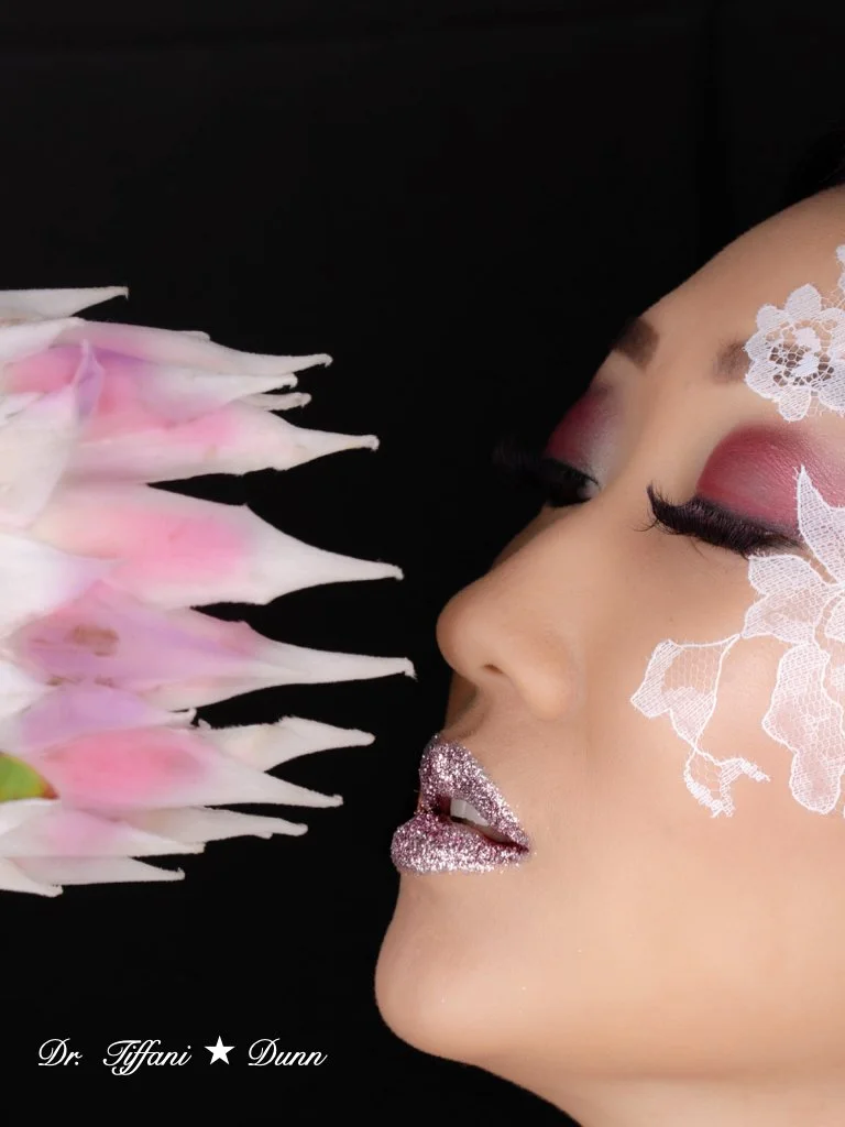 Close-up of a woman's face with pink makeup, glittery lips, and white lace face paint, with her eyes closed and nose nearly touching a cluster of pink and white flowers against a black background.
