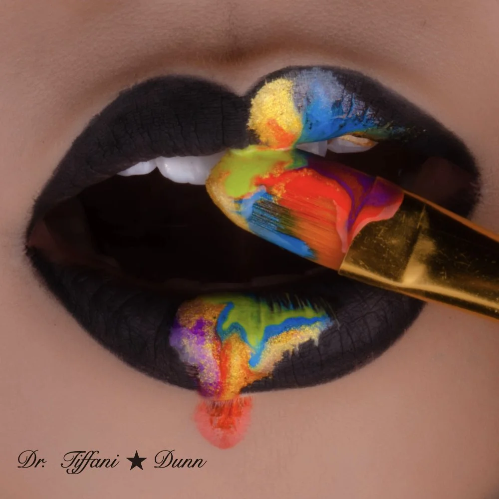Close-up of lips with black lipstick, colorful paint on the lips and tongue, and a gold paintbrush touching the tongue. The bottom left corner has text reading 'Dr. Tiffani Dunn'.