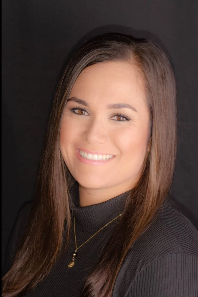 A woman with long brown hair smiling against a black background, wearing a black turtleneck and gold necklace.