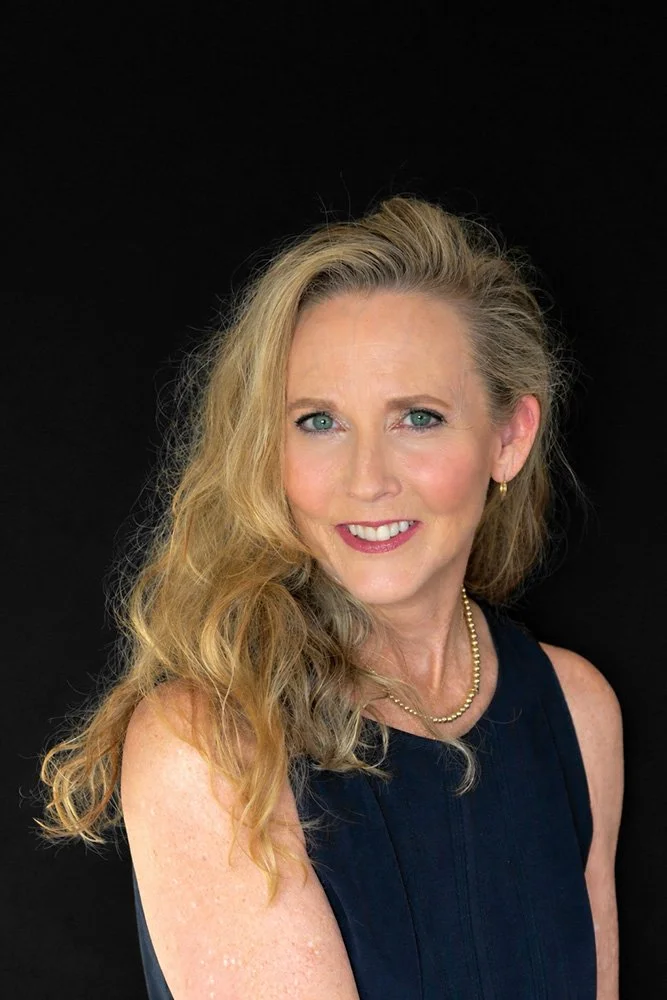 Portrait of a woman with long, wavy blonde hair, blue eyes, wearing a black sleeveless top, pearl necklace, and small earrings, smiling against a black background.