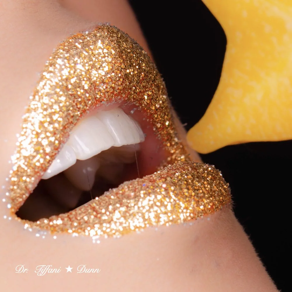 Close-up of a person's lips with gold glitter lipstick and gold glitter on the fingertips near the lips, with a yellow lipstick applicator in the background.