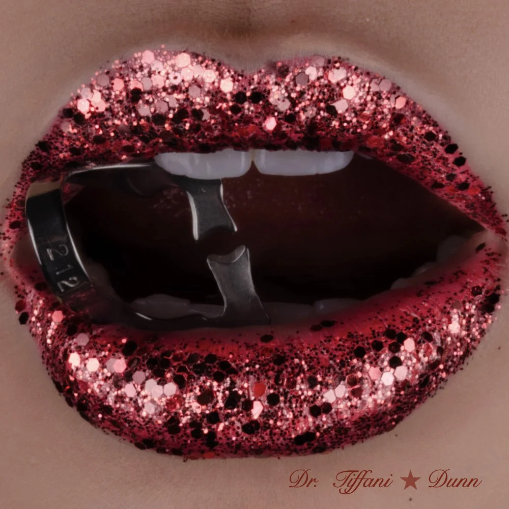 Close-up of lips with pink, red, and black glitter lipstick and dark lipstick inside the mouth, showing teeth and tongue with a black and silver wristwatch inside the mouth, with the signature 'Dr. Tiffani Dunn' at the bottom.