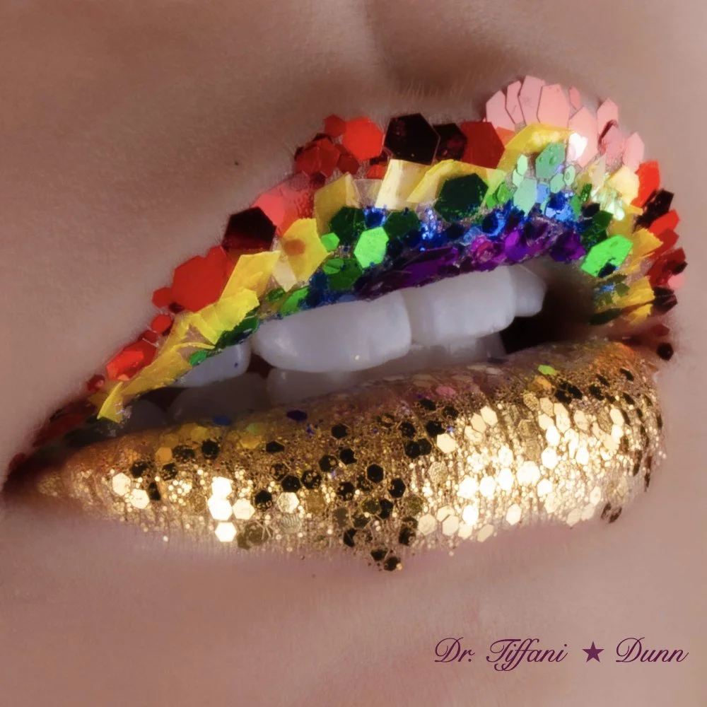 Close-up of lips with multicolored glitter makeup, featuring white teeth, green, purple, red, gold, and black glitter.