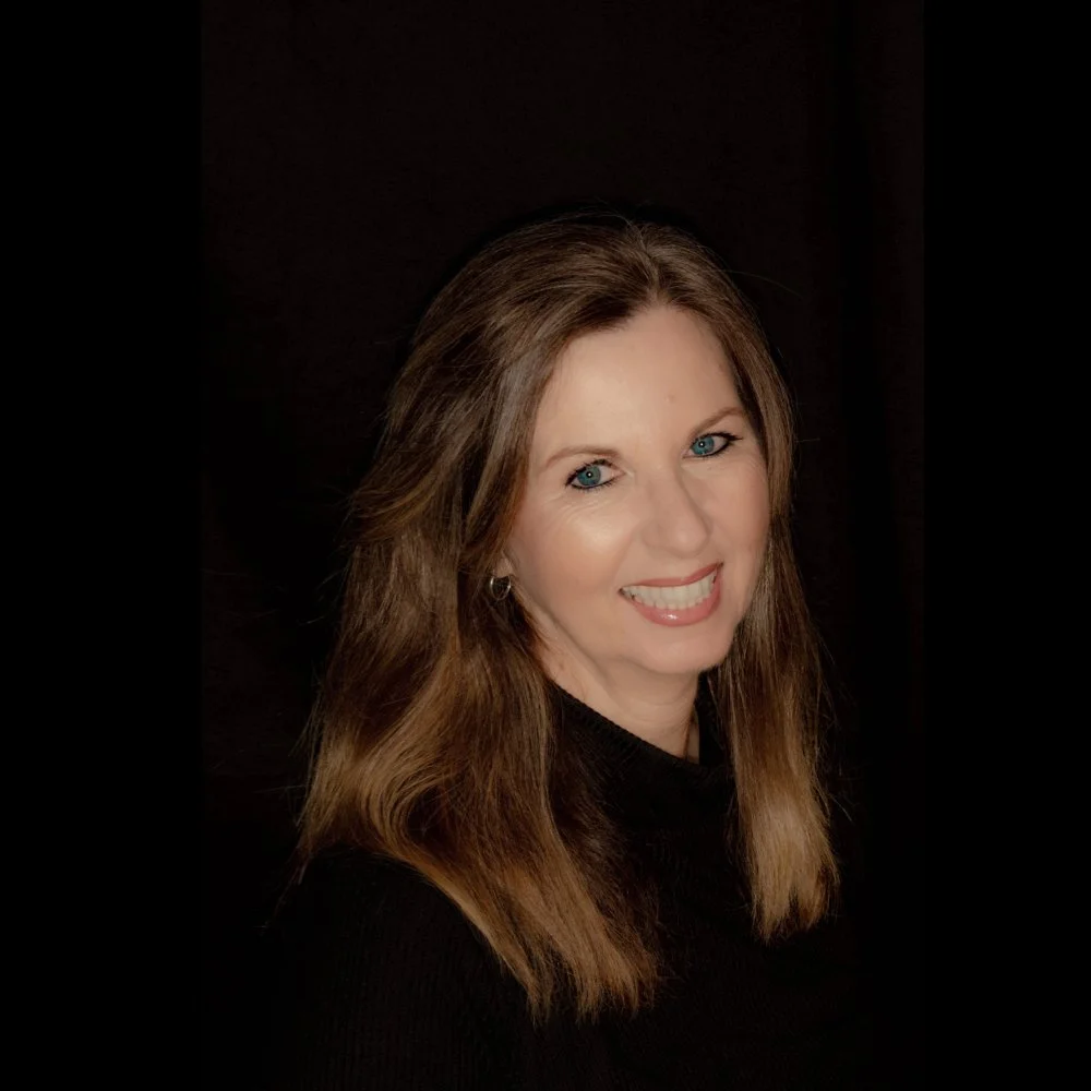 A woman with long brown hair and blue eyes smiling, wearing a black top, against a dark background.