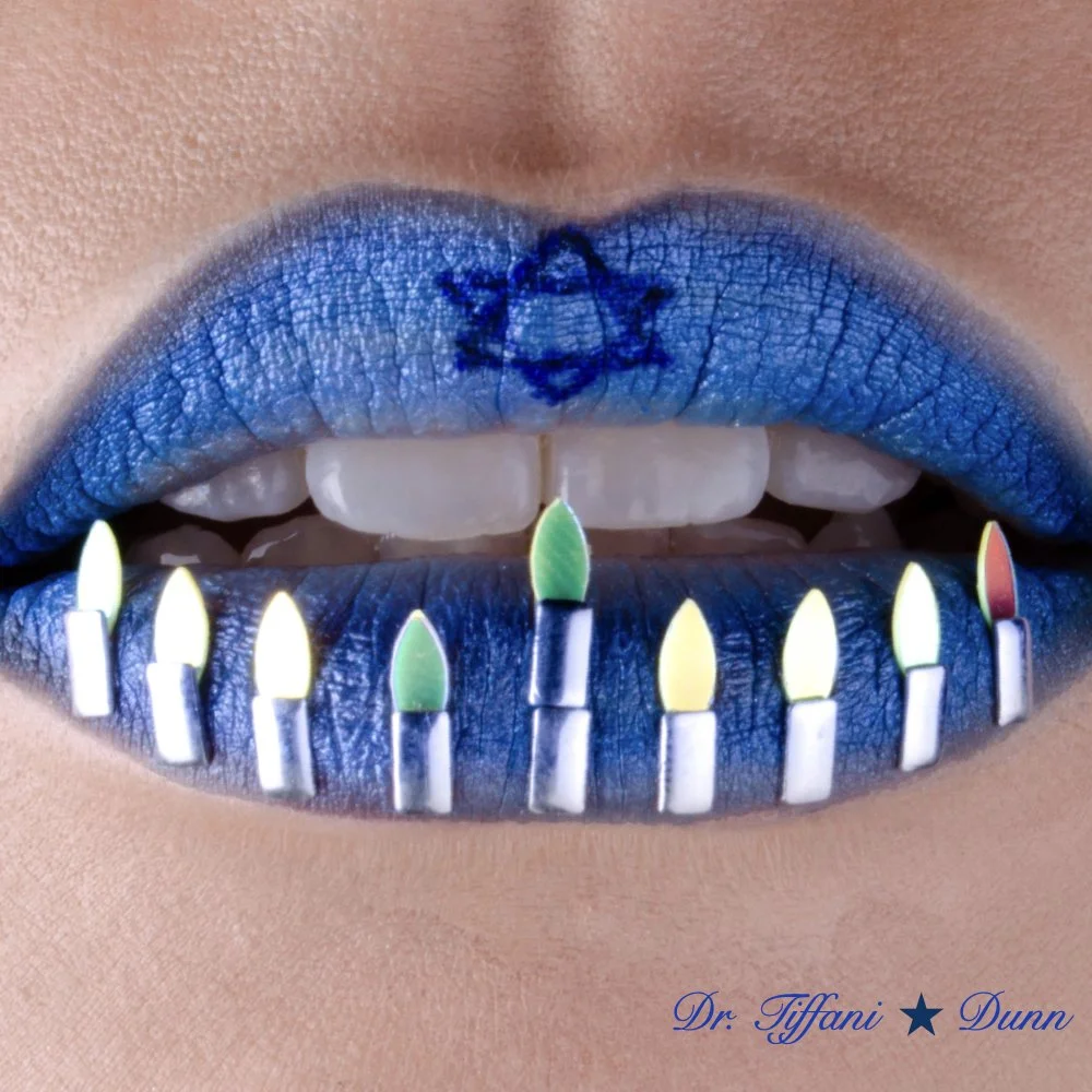 Close-up of lips with blue lipstick and a tattoo of a star on the upper lip. The teeth are visible, biting on a row of lit birthday candles on the lower lip. The candles are small with multicolored flames. The text "Dr. Tiffani Dunn" is at the bottom