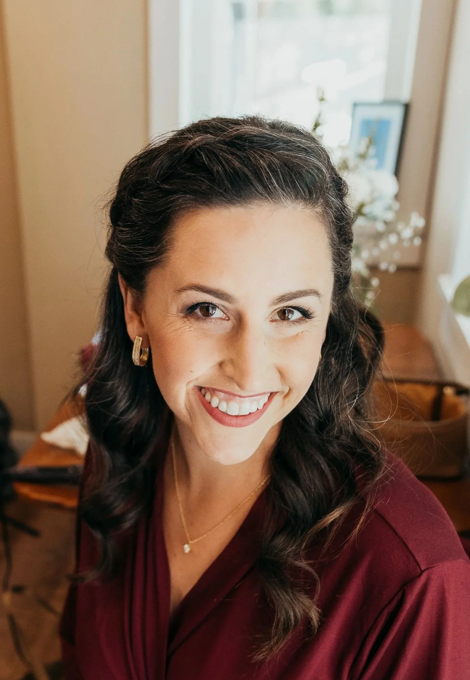 Close-up of a smiling woman with dark wavy hair, gold hoop earrings, a small necklace, and a maroon top indoors with a window and floral decorations in the background.