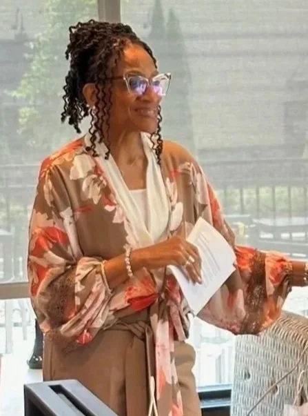 A woman with glasses and curly hair holds papers in a room with large windows and a balcony view.