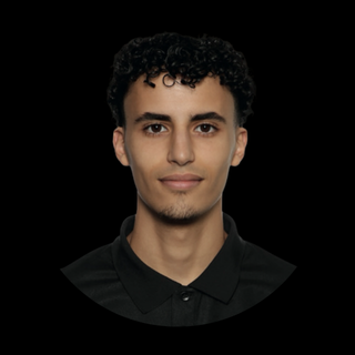 A young man with curly dark hair, wearing a black collared shirt, facing forward against a black background.